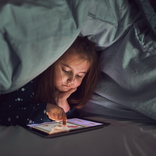 Little girl having fun watching, playing and listening to stories on tablet computer. Child having fun before bedtime laying under duvet using tablet